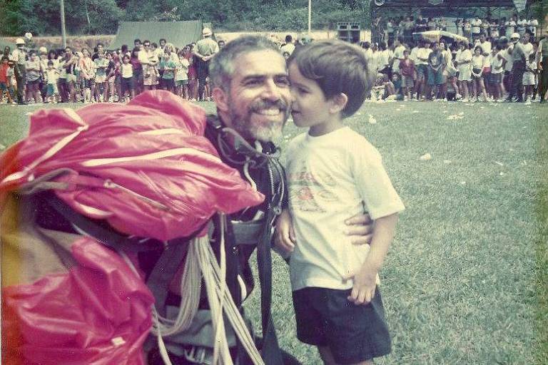 Homem de barba e farda, com paraquedas preso às costas, recebe beijo de criança