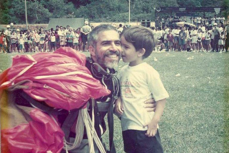 Homem de barba e farda, com paraquedas preso às costas, recebe beijo de criança