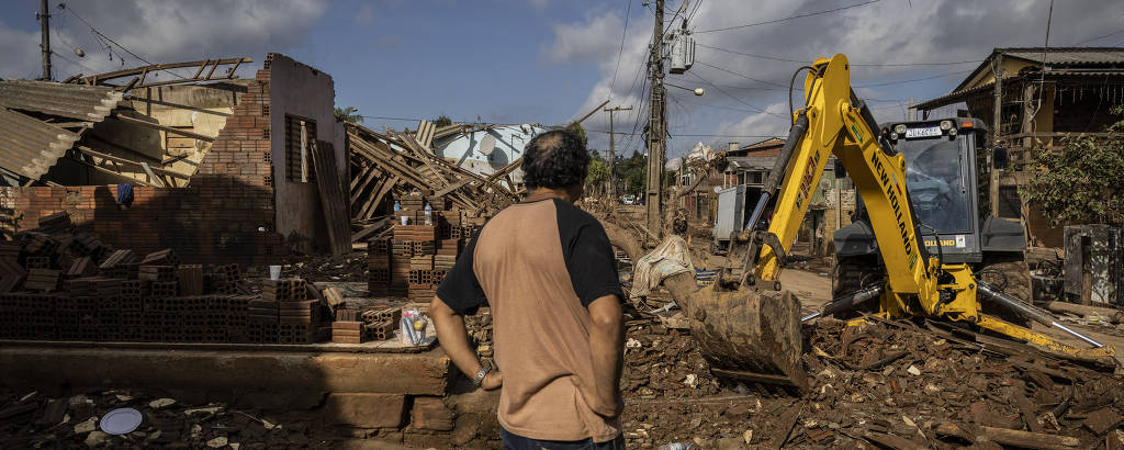 Um homem observa enquanto uma escavadeira trabalha entre os escombros de construções destruídas, sugerindo uma cena de demolição ou consequência de um desastre
