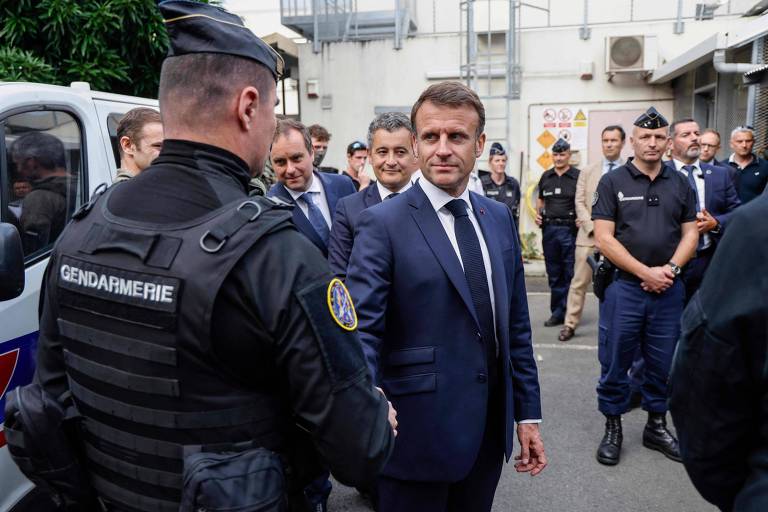 A esquerda, um policial pode ser visto de costas, usando colete e farda pretos, e uma boina cinza. Ele troca um aperto de mão com o Presidente Emannuel Macron que está de frente para a câmera usando um terno azul. Ao fundo, diversos policiais podem ser vistos observando o cumprimento, em frente à delegacia.