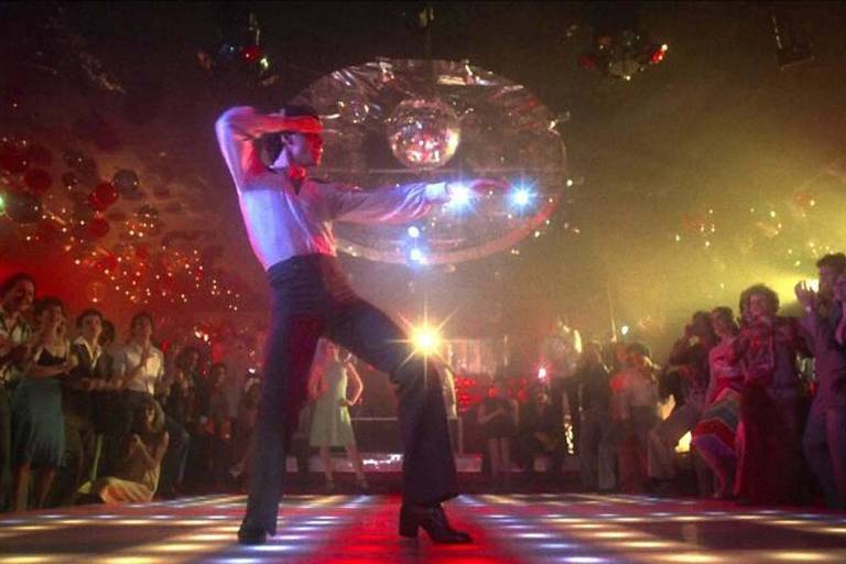 Um homem exibe seus movimentos de dança em uma pista de dança iluminada, cercado por uma multidão animada sob uma grande bola de discoteca. A atmosfera é eletrizante, com luzes refletindo no salão e balões vermelhos flutuando no ar, capturando a essência da era disco.