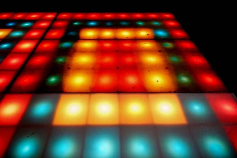 A imagem captura um piso iluminado por luzes coloridas, criando um mosaico vibrante de quadrados vermelhos, azuis, verdes e amarelos. A disposição das cores e a intensidade da luz conferem um efeito tridimensional, sugerindo movimento e energia.