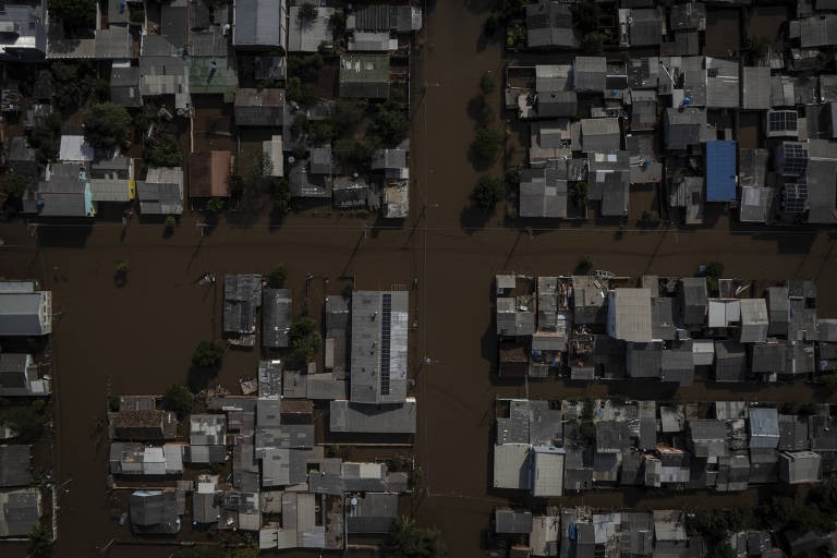 A imagem mostra uma vista aérea de uma área residencial densamente povoada, com várias casas parcialmente submersas em água de enchente. As ruas estão inundadas