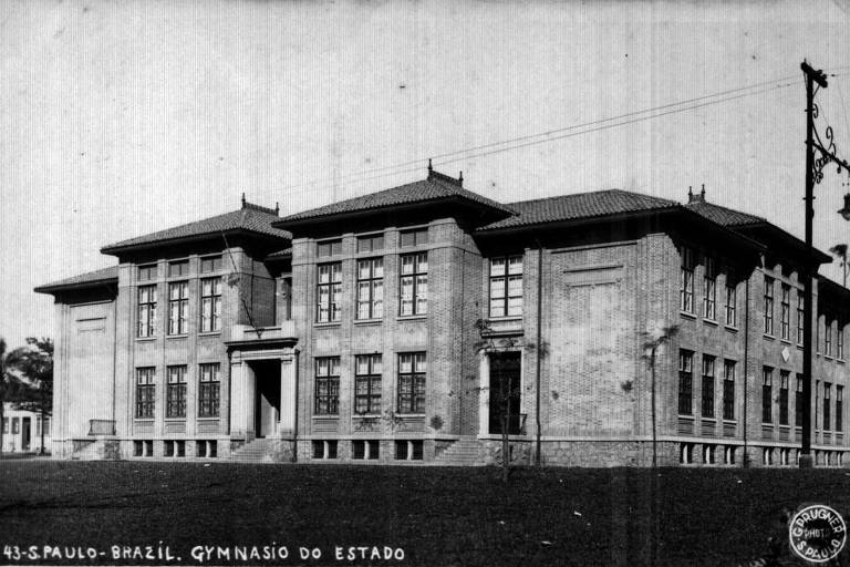 Edifício grande e simétrico com três pavimentos, janelas retangulares alinhadas e entrada central com escadaria. Fachada em estilo clássico, com telhado de várias águas e detalhes arquitetônicos simples. Foto em preto e branco, com céu claro ao fundo.
