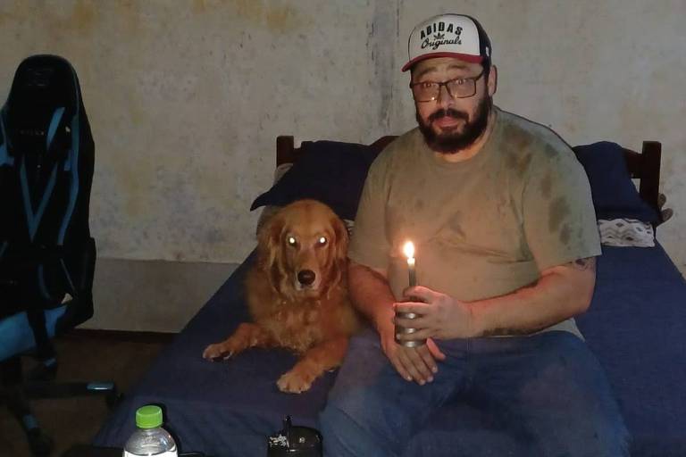 Homem branco sentado em um sofá, segurando uma vela com a mão, ao lado de um cachorro