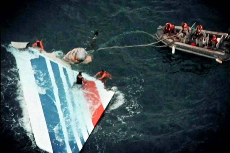 A imagem mostra uma operação de resgate em andamento no oceano, com várias pessoas usando coletes salva-vidas ao redor de um grande pedaço de destroços de avião, que tem as cores azul, branco e vermelho. Um bote inflável com resgatistas aproxima-se para ajudar os sobreviventes, destacando a gravidade e a urgência da situação