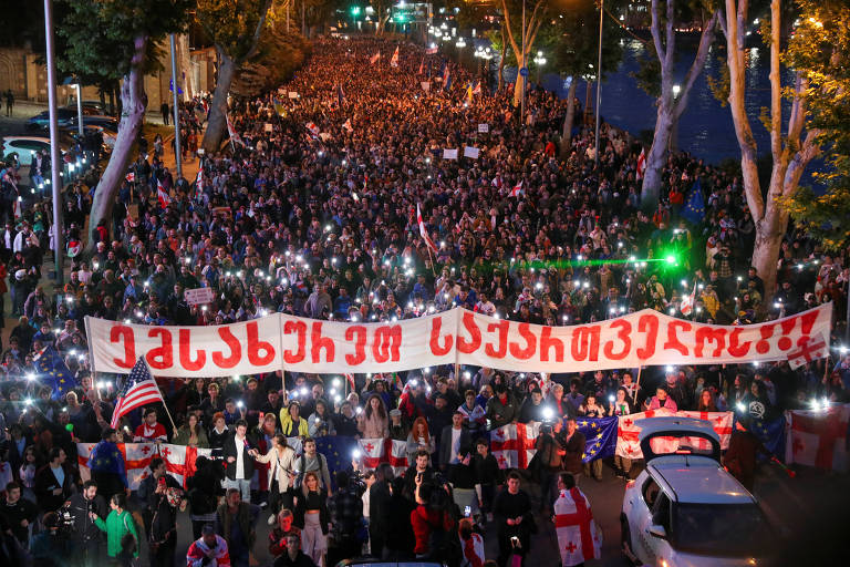 Uma multidão densa iluminada por luzes noturnas ocupa uma rua durante um protesto. Um grande banner com inscrições em um alfabeto não latino é carregado por manifestantes, enquanto bandeiras, incluindo uma dos Estados Unidos, são visíveis acima da multidão.