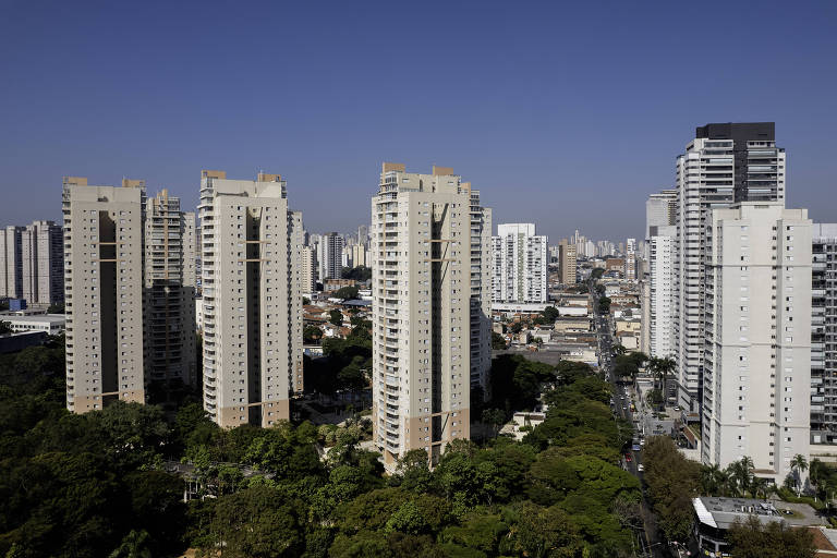Prédios do bairro Tatuapé, com fundo de céu azul sem nuvens; na porção abaixo da foto, o topo verde de árvores do parque Piqueri
