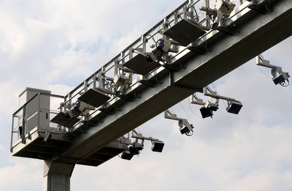 Estrutura de metal com câmeras e outros equipamentos forma um arco acima de uma rodovia, por onde passa um caminhão. Ao lado, há uma placa de sinalização. O céu está nublado.