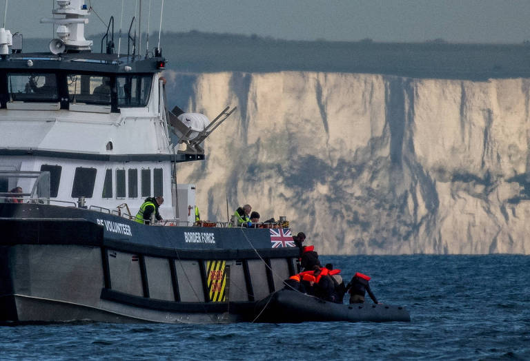 Um barco de patrulha navega próximo a um bote inflável com tripulantes vestidos com coletes salva-vidas vermelhos; ao fundo, há um penhasco