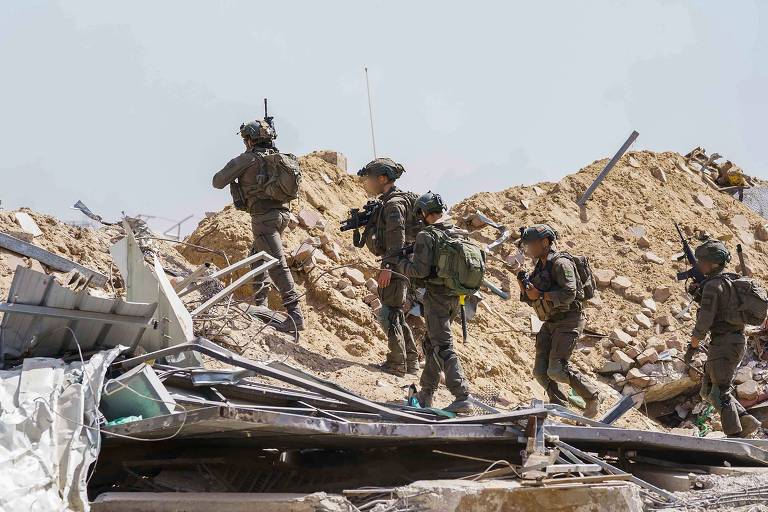 Cinco soldados equipados e armados patrulham uma área de escombros, com um deles posicionado em um ponto elevado
