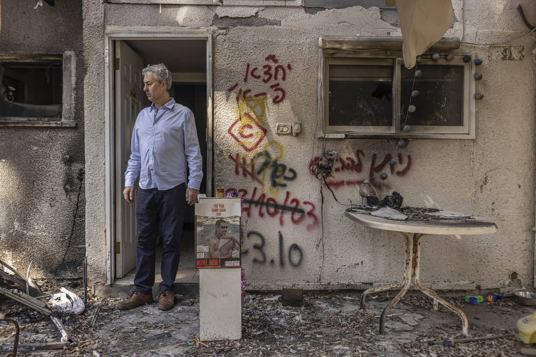 Homem parado na porta de um edifício danificado, com paredes chamuscadas e grafites. Ao seu lado, uma mesa com objetos queimados