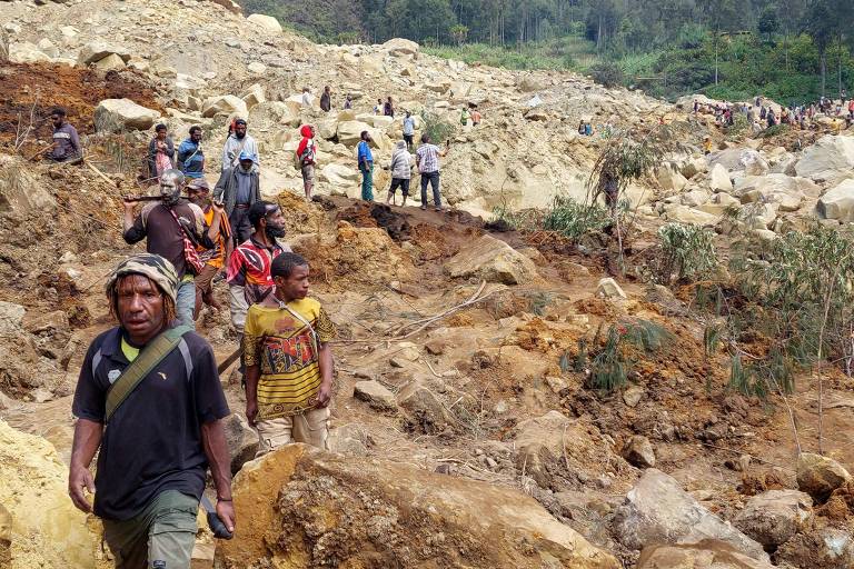 A imagem captura a luta de um grupo de pessoas para buscar sobreviventes em área onde houve soterramento de terra. Homens e mulheres, alguns carregando ferramentas, navegam por um terreno irregular e rochoso.
