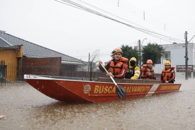 Quatro pessoas com capacetes laranjas e coloetes salva-vidas navegam em um barco numa inundação, onde se vê o nível da água de cor amarronzada a cima da altura de portas de construções, que são vistas ao fundo