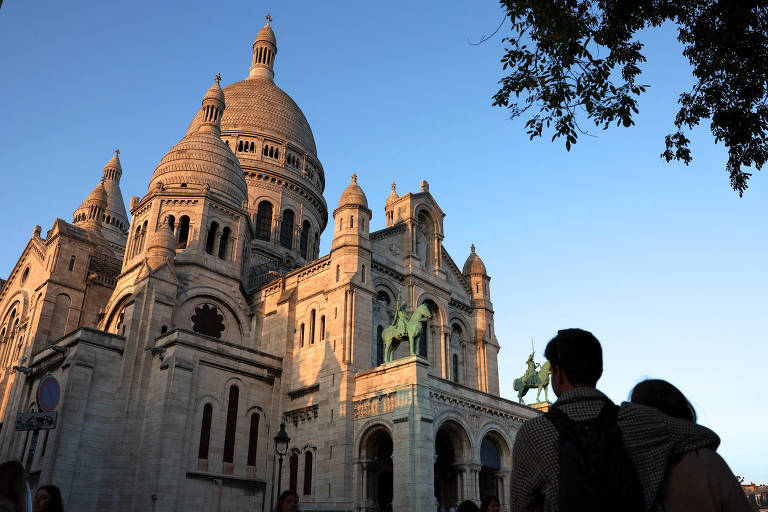 Entardecer na Basílica do Sagrado Coração em Paris, enquanto visitantes contemplam sua arquitetura. Há a silhueta de um casal, que está abraçado, em primeiro plano