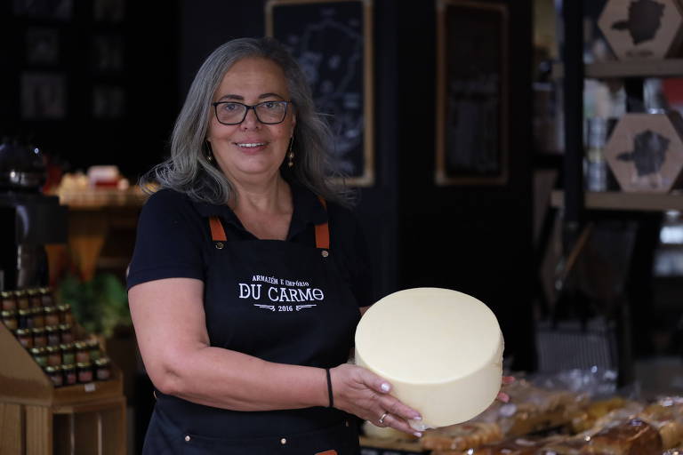 Uma mulher sorridente de meia idade, vestindo um avental preto com o nome "Empório Du Carmo" estampado, apresenta um grande queijo artesanal. Ela está em uma loja gourmet, cercada por produtos alimentícios, transmitindo uma atmosfera de tradição e qualidade