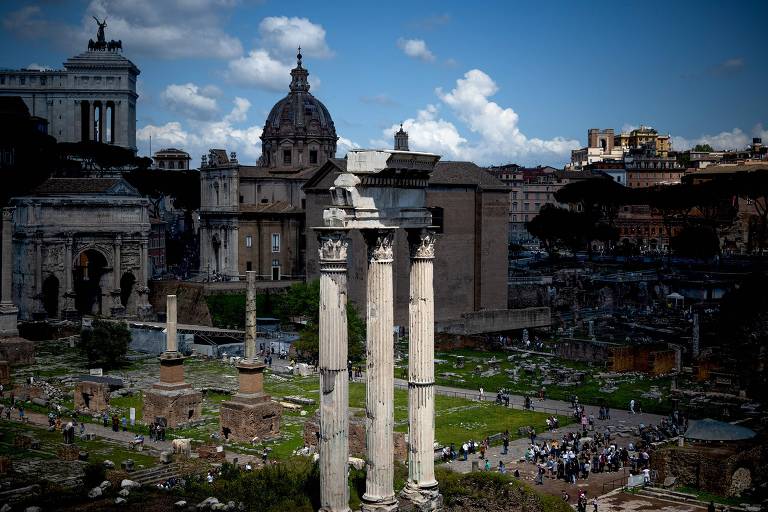 A imagem captura as colunas do Fórum Romano em primeiro plano, com visitantes explorando as antigas ruínas. Ao fundo, a arquitetura moderna da cidade se ergue