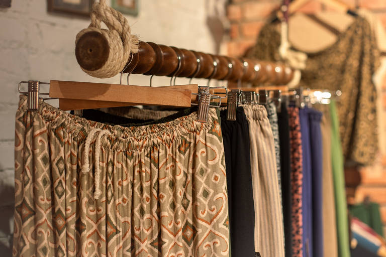 Uma seleção de calças com diferentes padrões e texturas está pendurada em um cabide de madeira, em um ambiente com decoração rústica e iluminação quente, sugerindo uma boutique de moda com um toque artesanal.