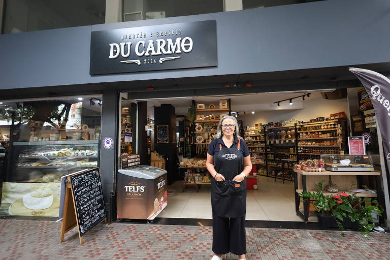 Uma mulher sorridente, vestindo um uniforme preto, está de pé em frente a loja "Empório Du Carmo". Há prateleiras visíveis e variedade de itens expostos na frente, incluindo vinhos e produtos frescos 