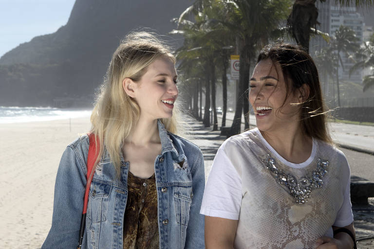Duas mulheres jovens compartilham um momento alegre sob o sol radiante, com uma praia deserta e palmeiras ao fundo, sugerindo um dia descontraído em um ambiente tropical.