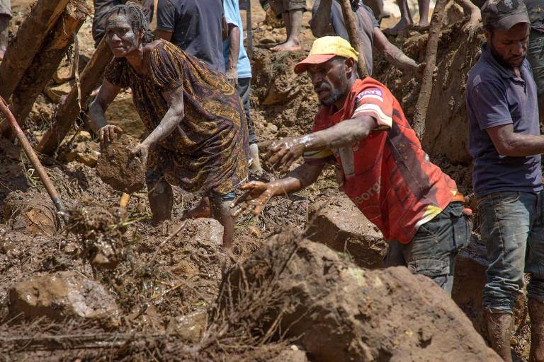 Mulher e homem de pele escura, cobertos em lama, usam pás e as mãos para mover rochas e restos do deslizamento que deixou vítimas