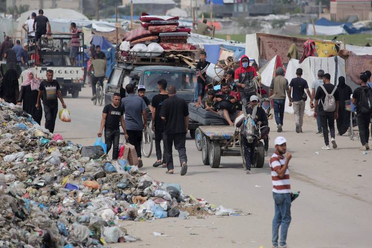 A imagem captura a agitação de uma rua movimentada, onde pessoas transitam entre barracas e veículos, com uma pilha de lixo em primeiro plano, destacando as condições desafiadoras do ambiente urbano que sofreu bombardeio.