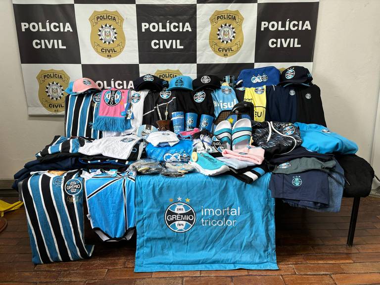 Mesa com vários produtos do Grêmio, como camisas, sapatos, chinelos e bonés estão organizados com fundo da Polícia Civil