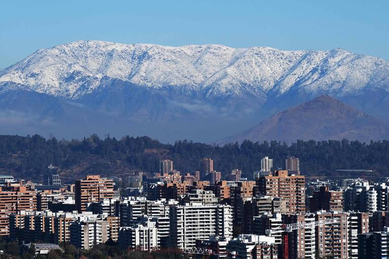 A imagem mostra uma paisagem urbana com vários prédios de diferentes alturas em primeiro plano, contrastando com as montanhas cobertas de neve ao fundo sob um céu claro e azul.
