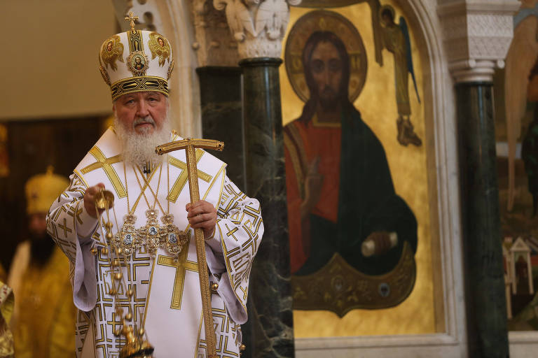 O patriarca ortodoxo russo está vestido com trajes cerimoniais, incluindo uma mitra dourada e um manto branco decorado, segura uma grande cruz enquanto conduz um serviço religioso. Ao fundo, uma pintura icônica de Jesus Cristo.