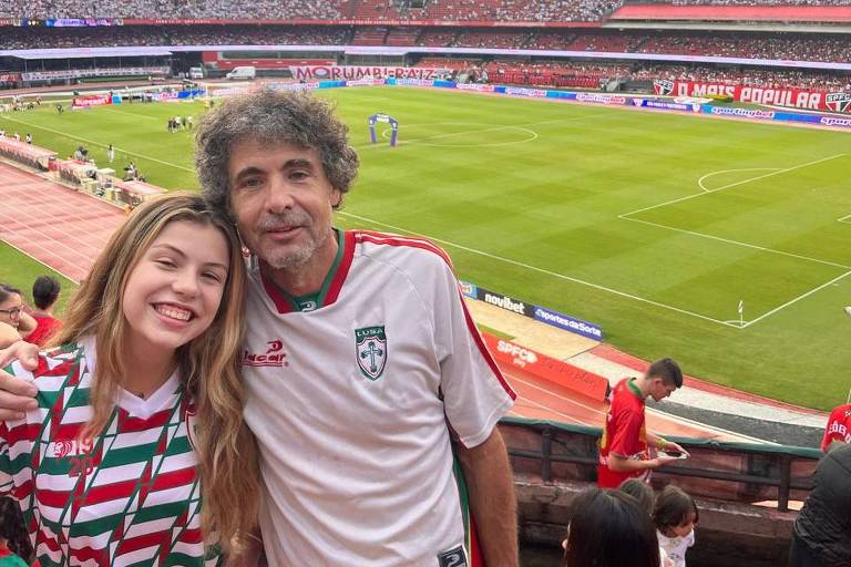 Paulo Vieira e sua filha em um jogo de futebol. Os dois estão abraçados e sorriem para a foto. Atrás, o campo de futebol.