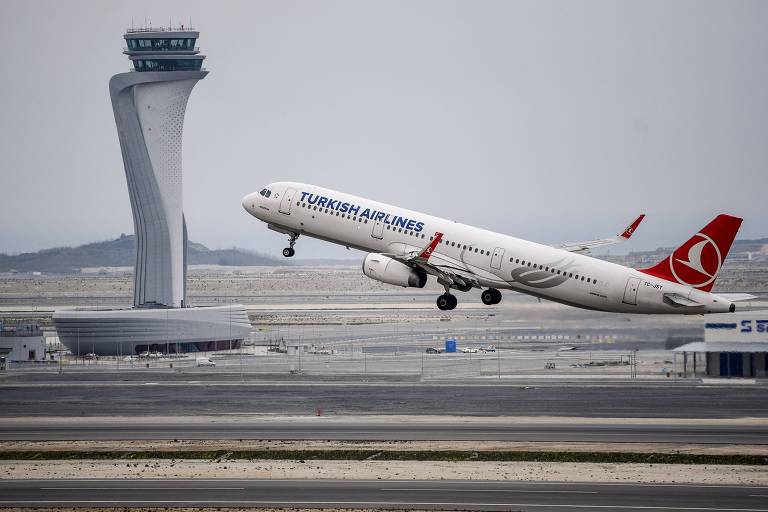 Um avião da Turkish Airlines levanta voo diante de uma torre de controle moderna e imponente, destacando-se contra um céu nublado e um horizonte de infraestrutura aeroportuária.