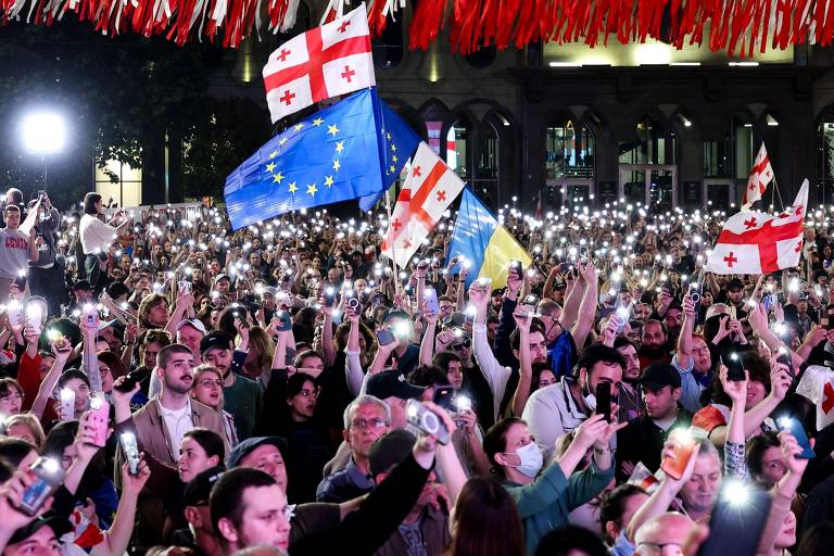 Uma multidão vibrante se reúne à noite, erguendo seus celulares iluminados em um ato de solidariedade ou protesto. Bandeiras da Geórgia e da União Europeia são agitadas com fervor, simbolizando um momento de união cívica e possivelmente um chamado por integração ou apoio internacional.