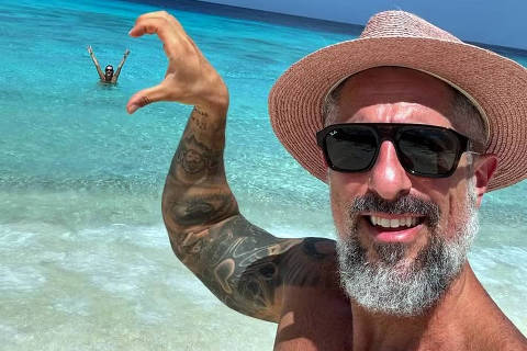 Marcos Mion em Curaçao *** Local Caption *** Um homem sorridente com chapéu e óculos escuros tira uma selfie na praia, posicionando sua mão de forma a parecer que está segurando uma pessoa ao fundo, que está no mar com os braços levantados, criando uma ilusão de ótica divertida.