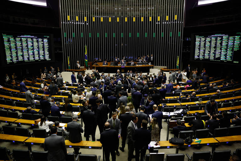 A imagem captura uma sessão noturna em uma câmara legislativa, com políticos reunidos no plenário. As luzes focam no centro, onde alguns membros estão em pé, possivelmente em um debate ou discussão. 