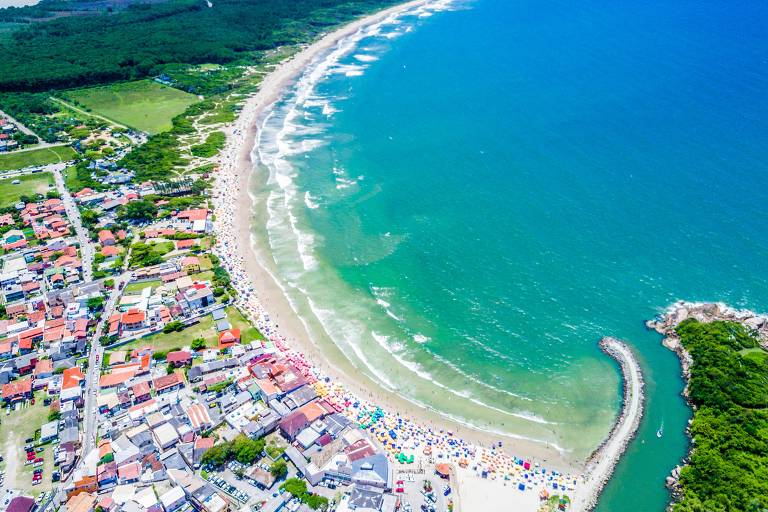 Vista aérea de praia catarinense, onde as águas cristalinas encontram a areia branca, delineando a curvatura da costa. A paisagem é pontuada por casas com telhados coloridos e vegetação verdejante 
