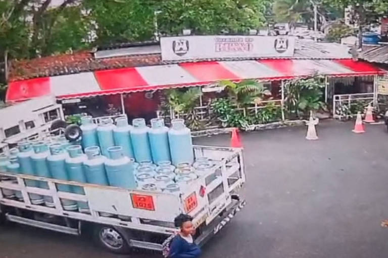 Um caminhão de entrega está estacionado ao lado de uma estrada, carregado com numerosos botijões de gás azuis. Dois homens parecem estar trabalhando; um está caminhando em direção à traseira do veículo, enquanto o outro está próximo ao caminhão, em movimento