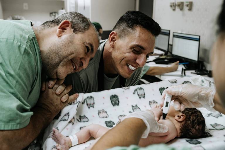 Dois homens exibem sorrisos radiantes enquanto se inclinam amorosamente sobre um recém-nascido que está deitado em uma cama de hospital. Um deles, vestindo luvas, segura um termômetro, indicando que estão cuidando da saúde do bebê. O ambiente sugere que estão em uma sala de parto ou unidade neonatal, compartilhando um momento de ternura e felicidade após o nascimento.