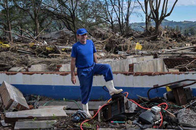 Um homem em uniforme azul está parado sobre os escombros de uma construção destruída, olhando para o chão. Ele está cercado por destroços, incluindo pedaços de concreto, cabos e equipamentos danificados, com árvores e um céu claro ao fundo.