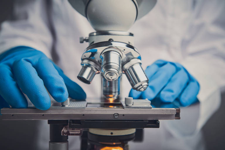 Um cientista em um laboratório, vestindo um jaleco branco e luvas azuis, ajusta um microscópio óptico , preparando-se para examinar uma amostra não visível na imagem
