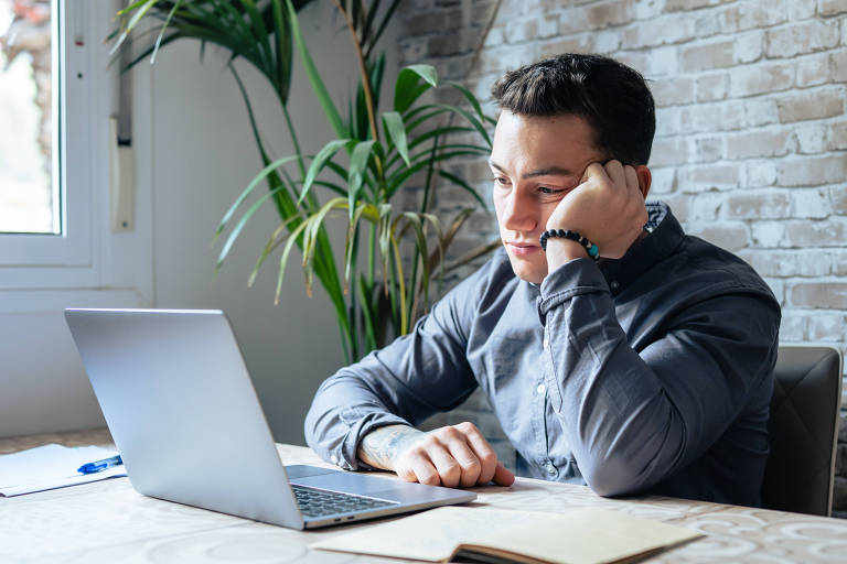 Um homem vestindo uma camisa cinza está concentrado em seu trabalho enquanto usa um laptop em um escritório em casa bem iluminado. Ele apoia o rosto na mão, sugerindo reflexão ou leitura atenta. Ao fundo, uma janela deixa entrar luz natural e uma planta adiciona um toque de verde ao ambiente.