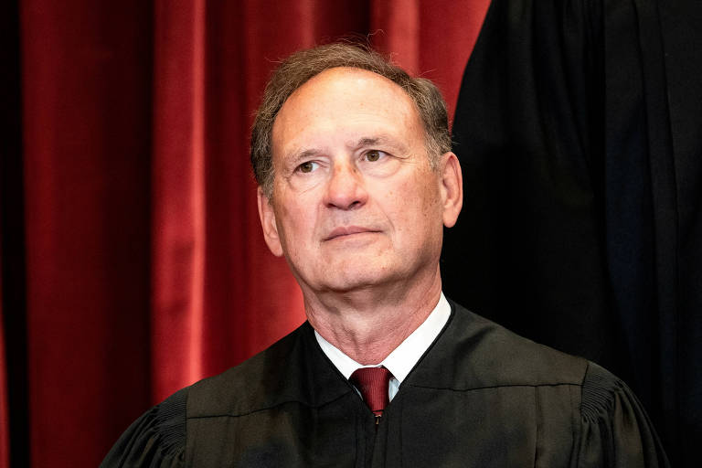 Um homem branco vestindo uma toga de juiz posa com uma expressão séria em frente a uma cortina vermelha