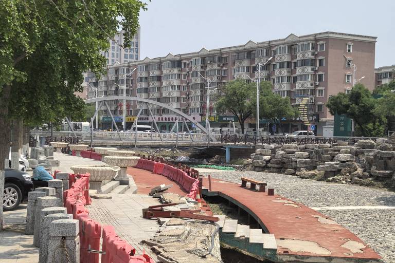 A imagem mostra um canteiro de obras ao longo de uma calçada, com foco em uma estrutura de tijolos vermelhos parcialmente construída. Ao fundo, há um prédio de apartamentos de vários andares e uma ponte de pedestres que atravessa um canal ou rio