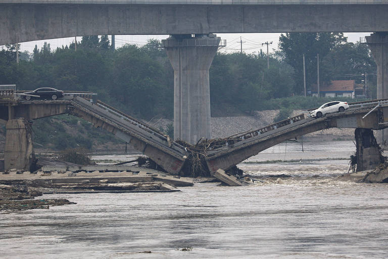 A imagem mostra uma ponte parcialmente destruída sobre um rio, com suas estruturas de concreto expostas e danificadas. Dois veículos estão parados perigosamente perto da borda do trecho quebrado