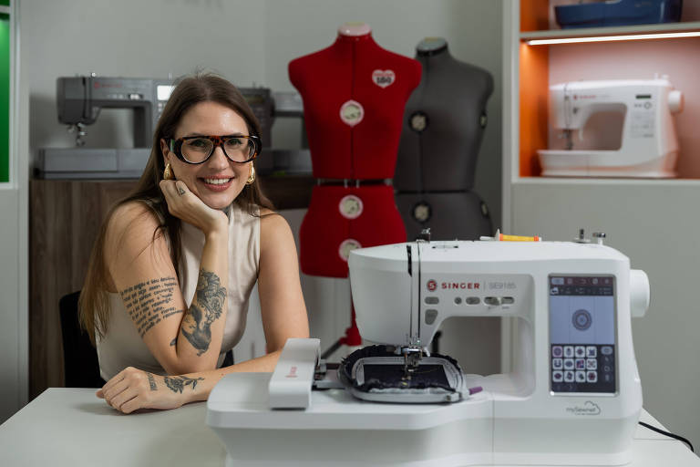 Mulher branca, jovem de óculos e tatuagem, cabelo longo castanho, ao lado de máquina de costura branca, com manequim vermelho ao fundo
