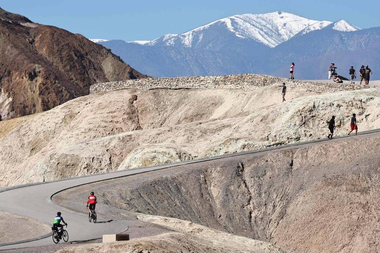 Foto horizontal mostra paisagem montanhosa árida com pessoas caminhando e pedalando na estrada de asfalto que corta o morro