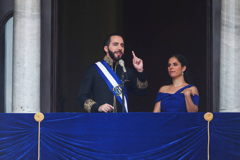 Vestido com um terno e faixa presidencial, Nayib Bukele discursa ao lado de sua esposa, Gabriela de Bukele, vestida de azul, na sacada do Palácio Nacional


