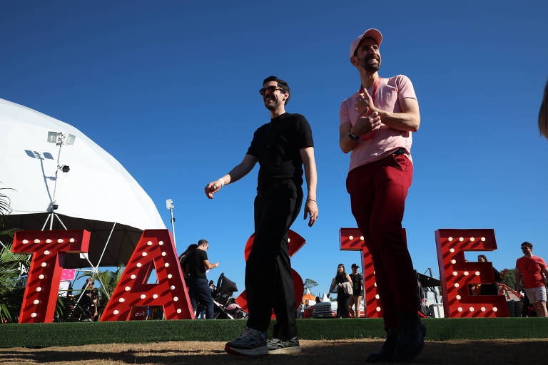 Dois homens caminham sorridentes sob um céu azul límpido, passando por uma instalação artística com a palavra "TASTE" em letras vermelhas iluminadas. O ambiente mostra um evento ao ar livre