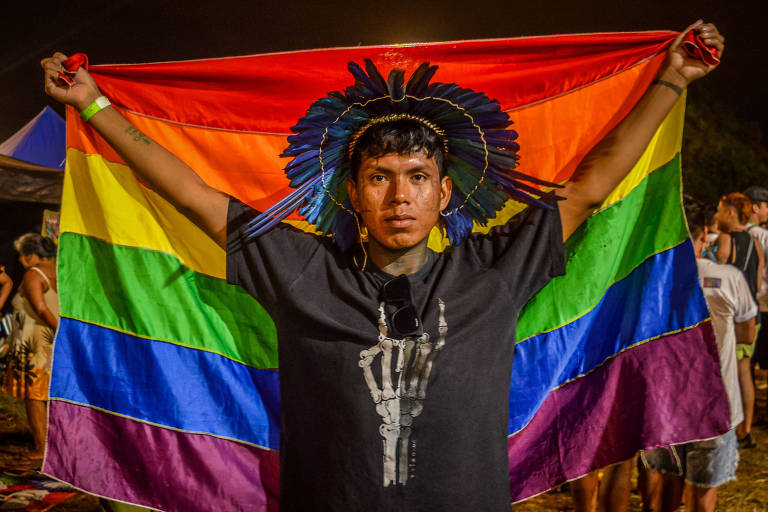 Um rapaz indígena com cocar colorido na cabeça, segura a bandeira do arco-íris, símbolo do orgulho LGBTQIA+. Ele usa uma camisa preta. No fundo, o céu noturno. 