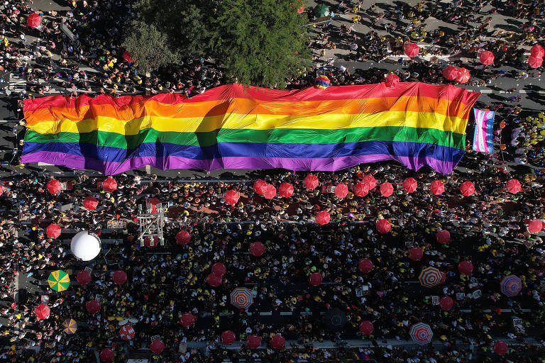 A imagem mostra uma grande multidão reunida em uma celebração do orgulho LGBTQ+， com um enorme banner de arco-íris estendido sobre a multidão. O fundo é preenchido com pessoas e balões vermelhos， enquanto algumas árvores são visíveis na parte superior da imagem.