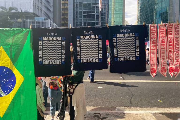 A imagem mostra três camisas pretas iguais estampadas com a lista de músicas da Madonna que ela canta nos shows. As camisas estão penduradas em uma corda e estão ao lado de uma bandeira do Brasil. No fundo alguns prédios da Avenida Paulista.